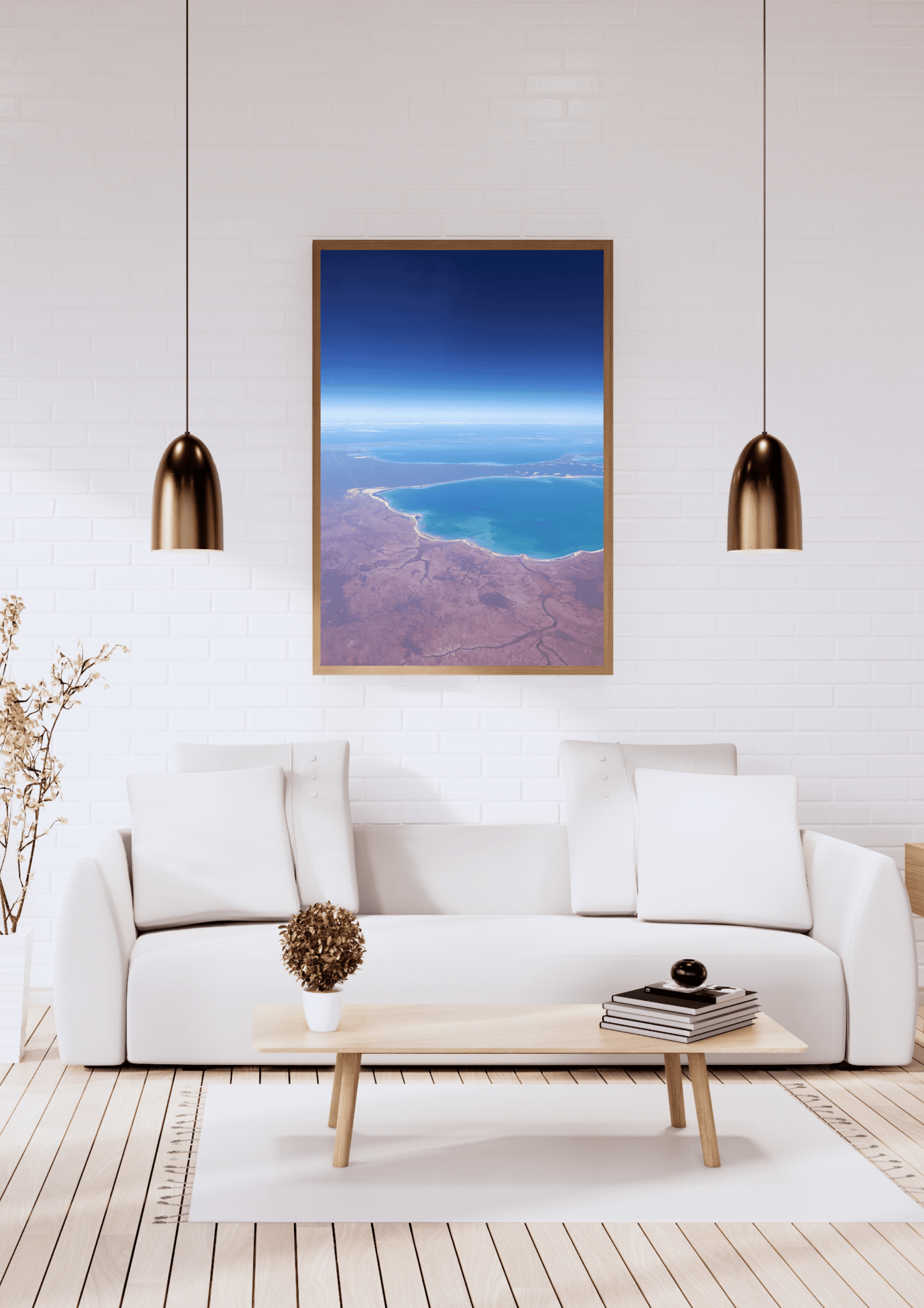 A framed art print depicting an aerial view of the deeply contrasting deep blue ocean against the red earth along the Western Australian coastline, displayed on a white brick wall above a modern white sofa in a bright living room.