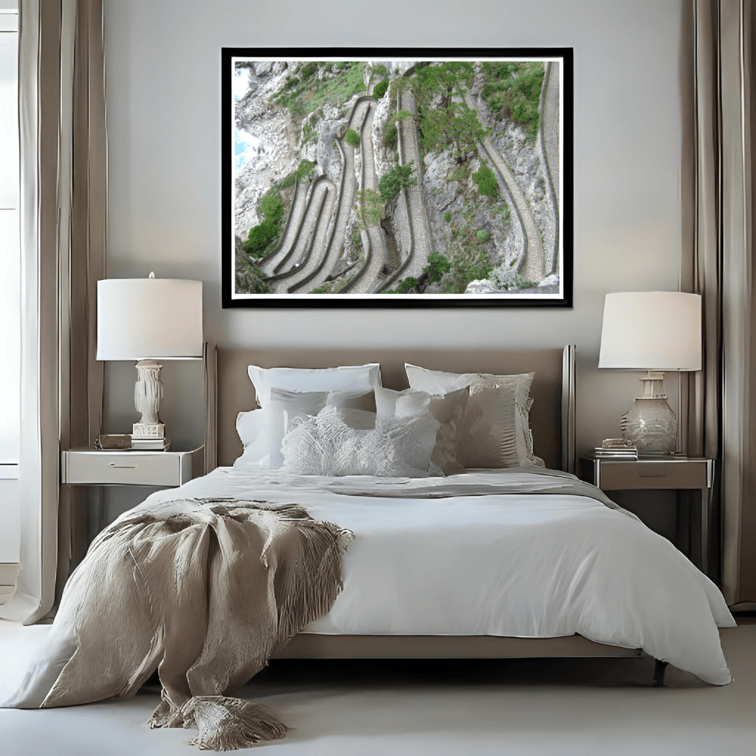 A framed wall art print depicting a winding paved path in Capri, displayed above a neatly made bed in a modern, neutral-toned bedroom.