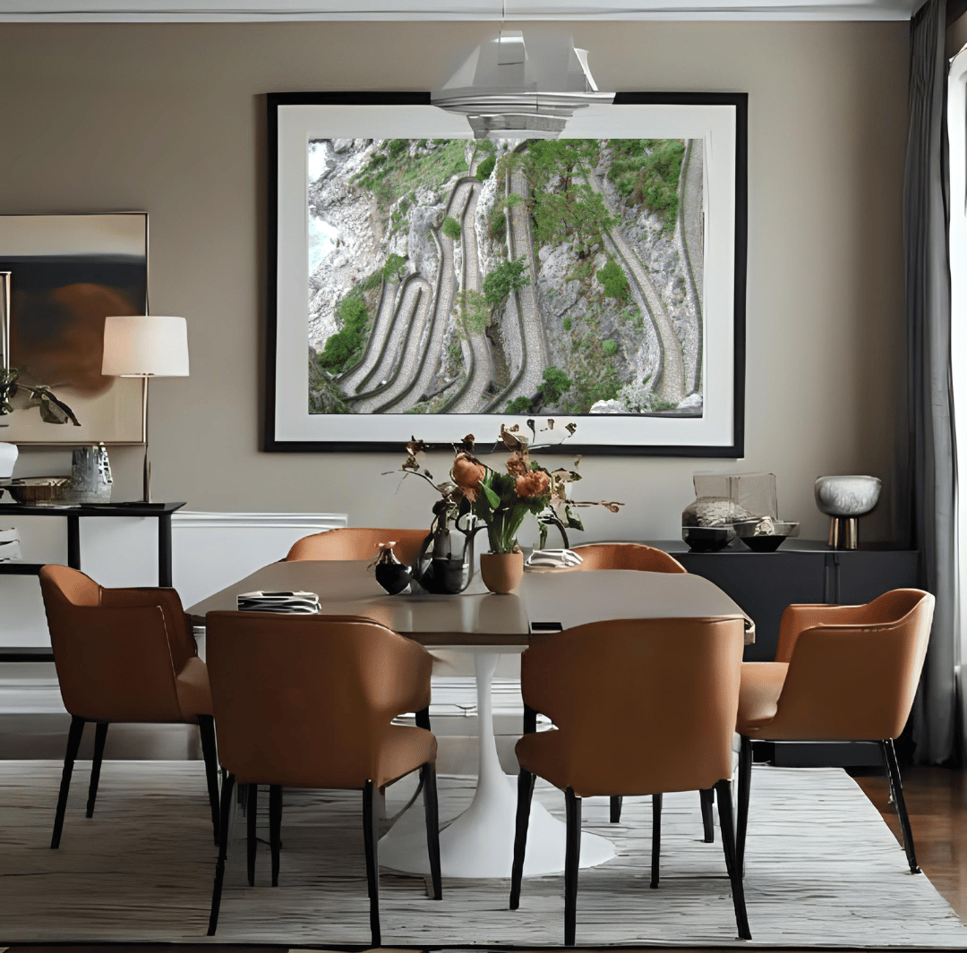 A large framed art print depicting a winding winding paved path in Capri hangs on a taupe wall in a modern dining room setting with brown upholstered chairs and a white pedestal table.