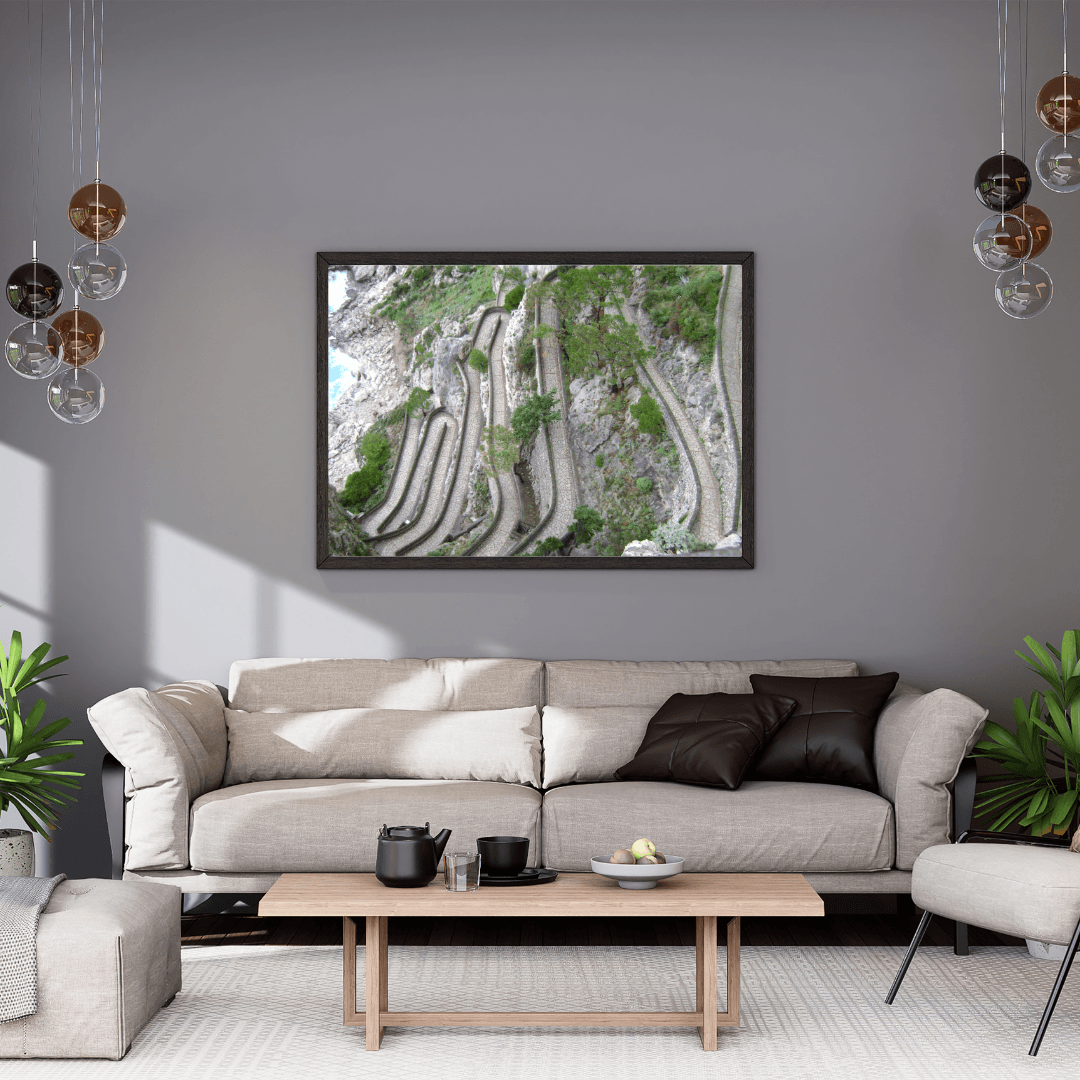 A framed wall art print depicting winding winding paved path in Capri through a green and gray rocky landscape, displayed in a modern living room setting above a light beige sofa.