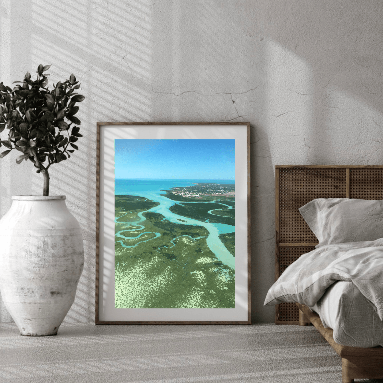 A framed landscape art print depicting an aerial view of the winding crystal blue waterways of the ocean through the green bushland, styled in a bedroom setting next to a bed and a white vase with a plant.