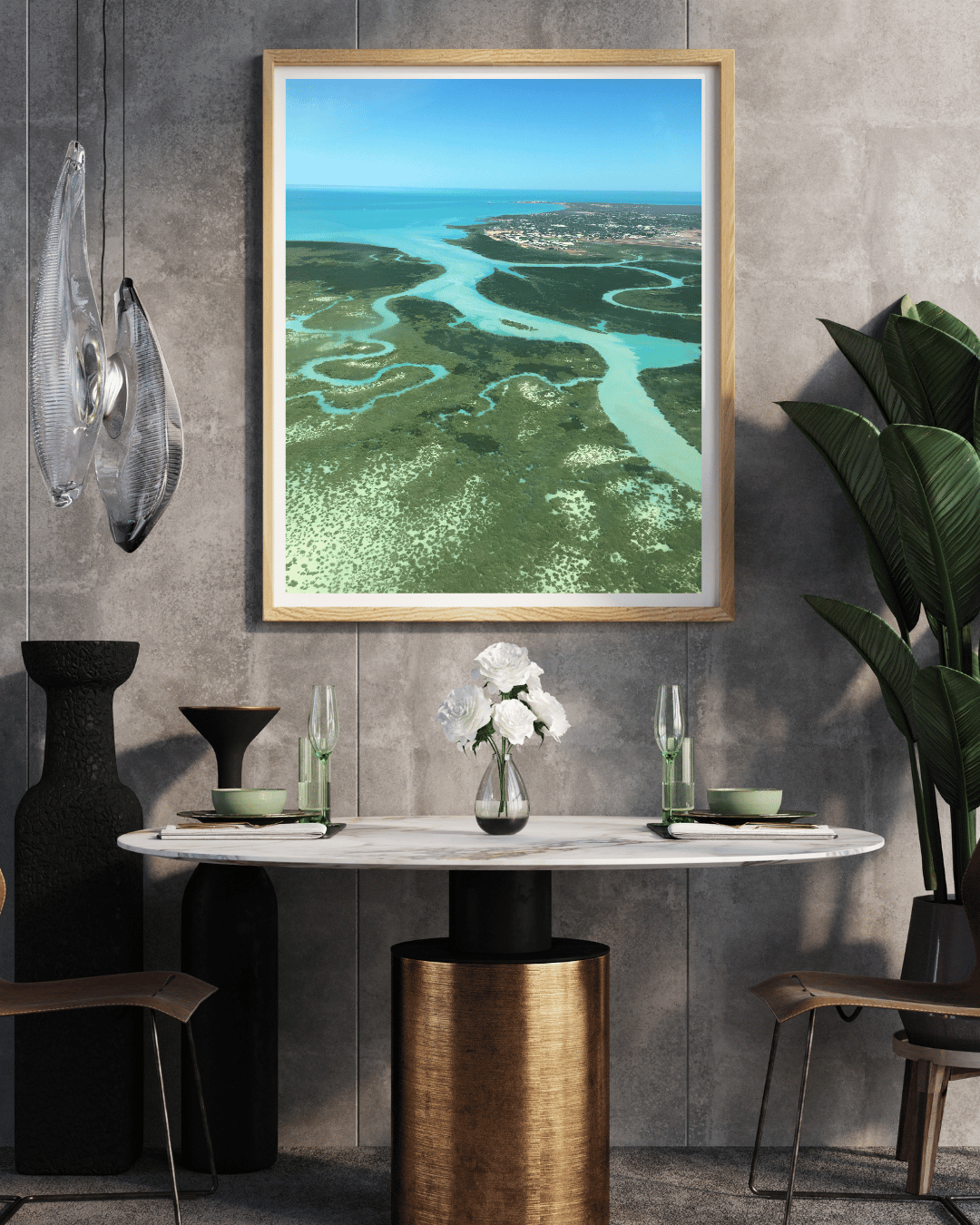 A framed aerial landscape photograph of an aerial view of the winding crystal blue waterways of the ocean through the green bushland, displayed on a gray concrete wall above a dining table in a modern styled room.