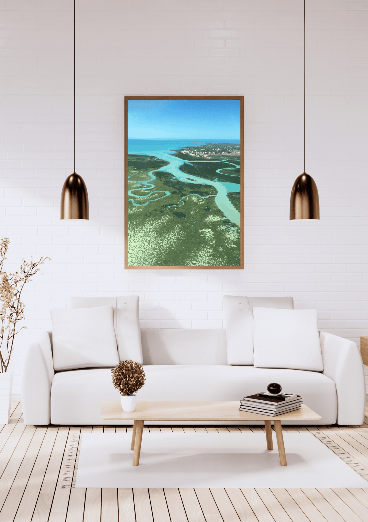 A framed aerial landscape photograph of an aerial view of the winding crystal blue waterways of the ocean through the green bushland, displayed on a white brick wall above a modern white sofa in a bright living room.