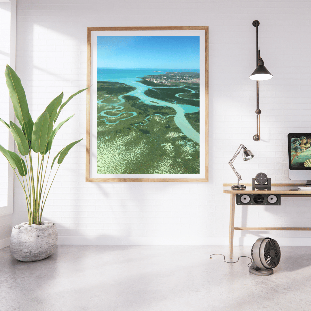 A framed landscape art print depicting an aerial view of the winding crystal blue waterways of the ocean through the green bushland, displayed on a white wall in a modern, sunlit room with a plant, desk, and lamp.
