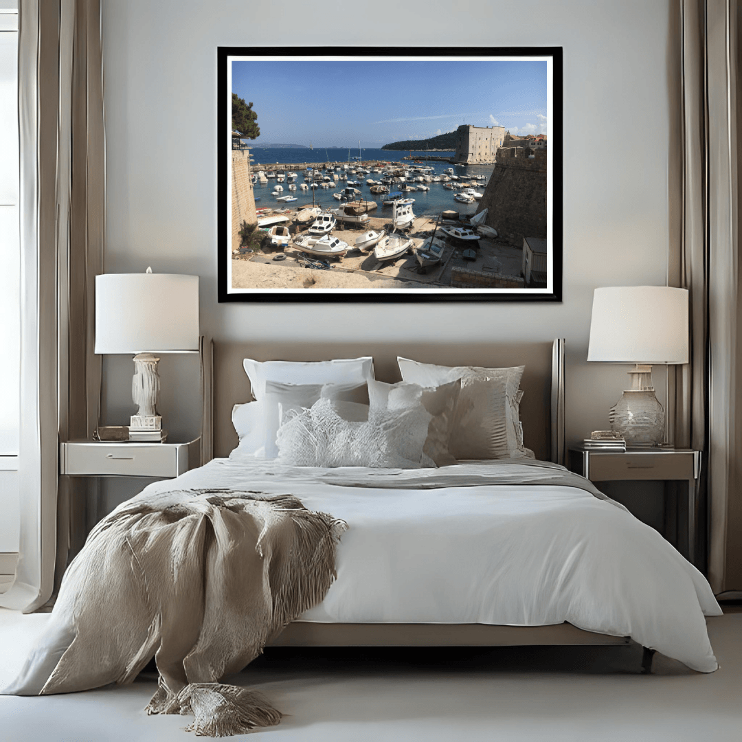 A framed art print depicting a coastal harbor scene with boats and stone buildings in Dubrovnik, displayed on a neutral wall above a modern bed in a styled bedroom setting.