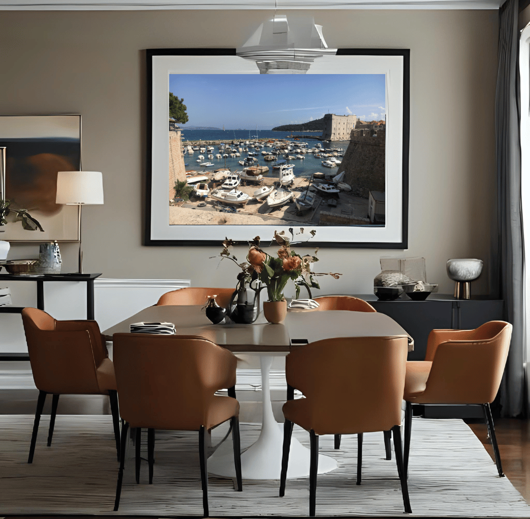 A large framed art print depicting a coastal harbor scene with boats and stone buildings in Du, displayed on a beige wall in a modern dining room setting with a dining table and six brown chairs.rovnik
