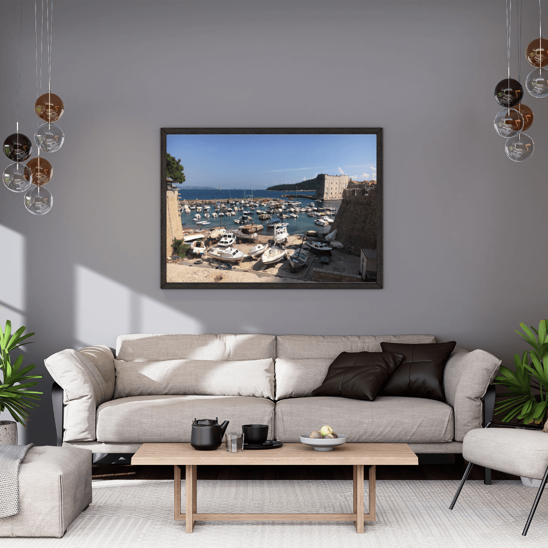 A framed art print depicting a scenic harbor with boats and a stone wall in Dubrovnik, displayed on a gray wall above a modern beige sofa in a styled living room.