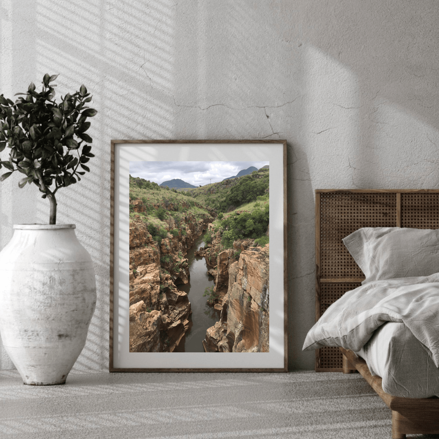 A framed landscape art print of a deep canyon with a river, styled in a modern bedroom setting next to a bed and a white vase with a plant.