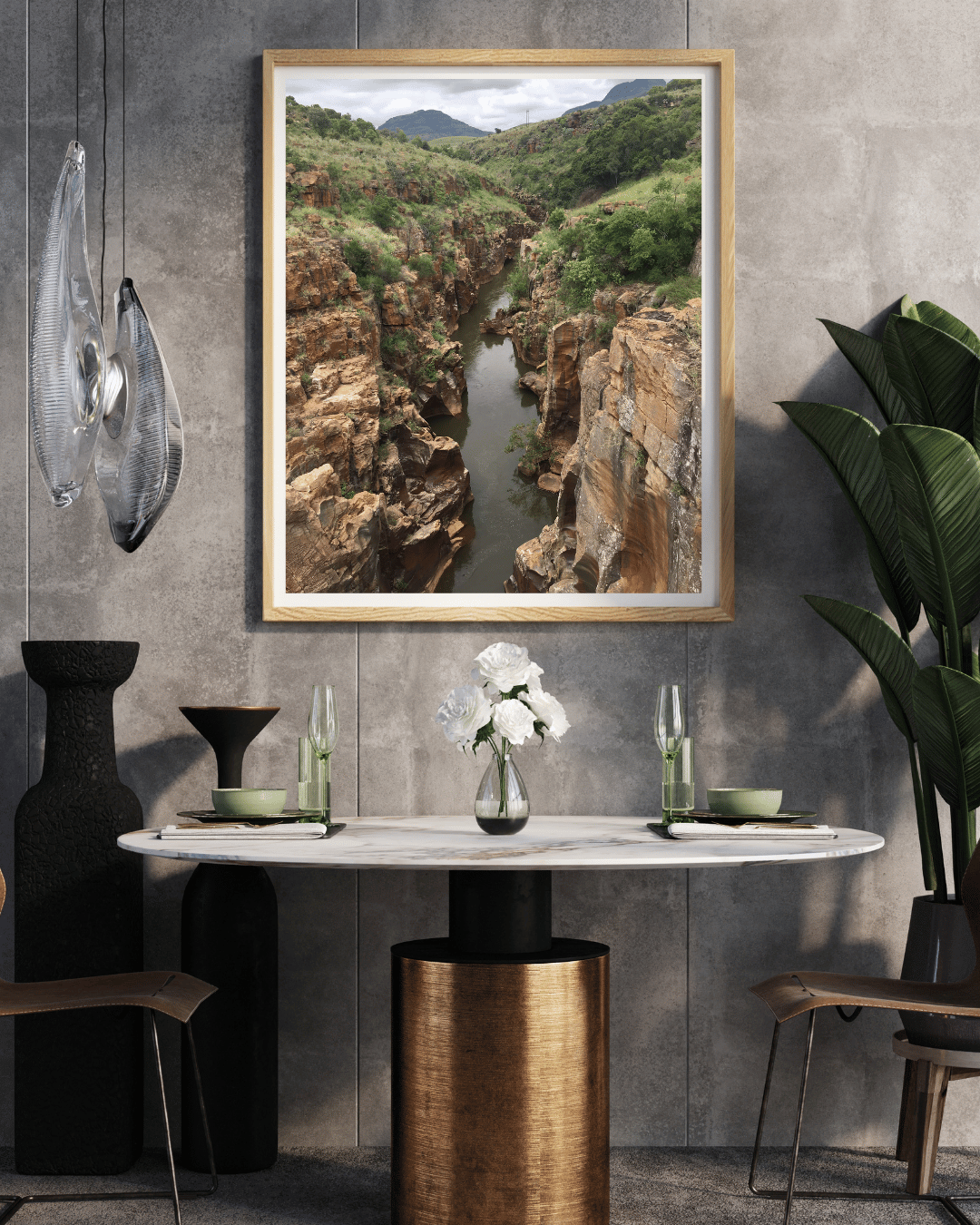 A framed landscape art print depicting a deep rocky canyon with a river flowing through it, hung on a textured gray wall above a dining table in a modern styled room.