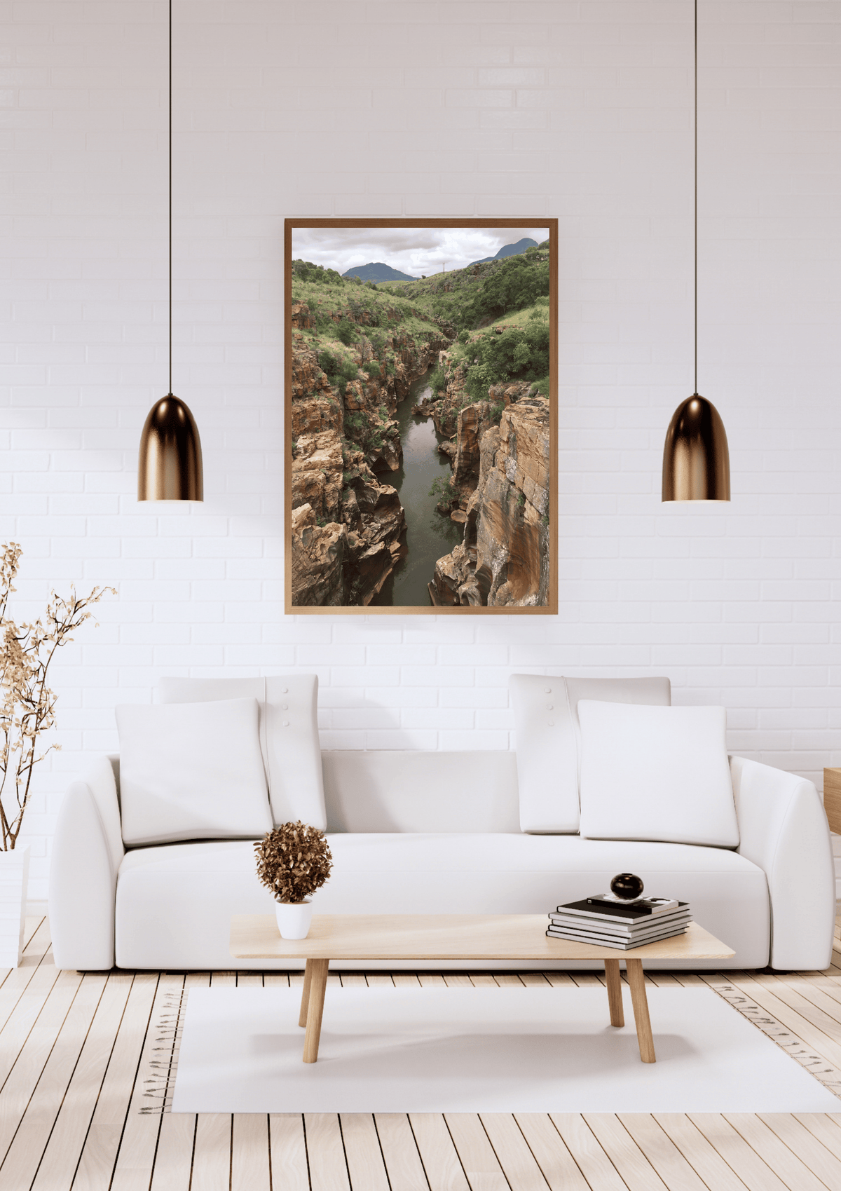 A framed landscape art print of a deep rocky canyon with a river flowing through it, displayed on a white brick wall above a modern white sofa in a styled living room.