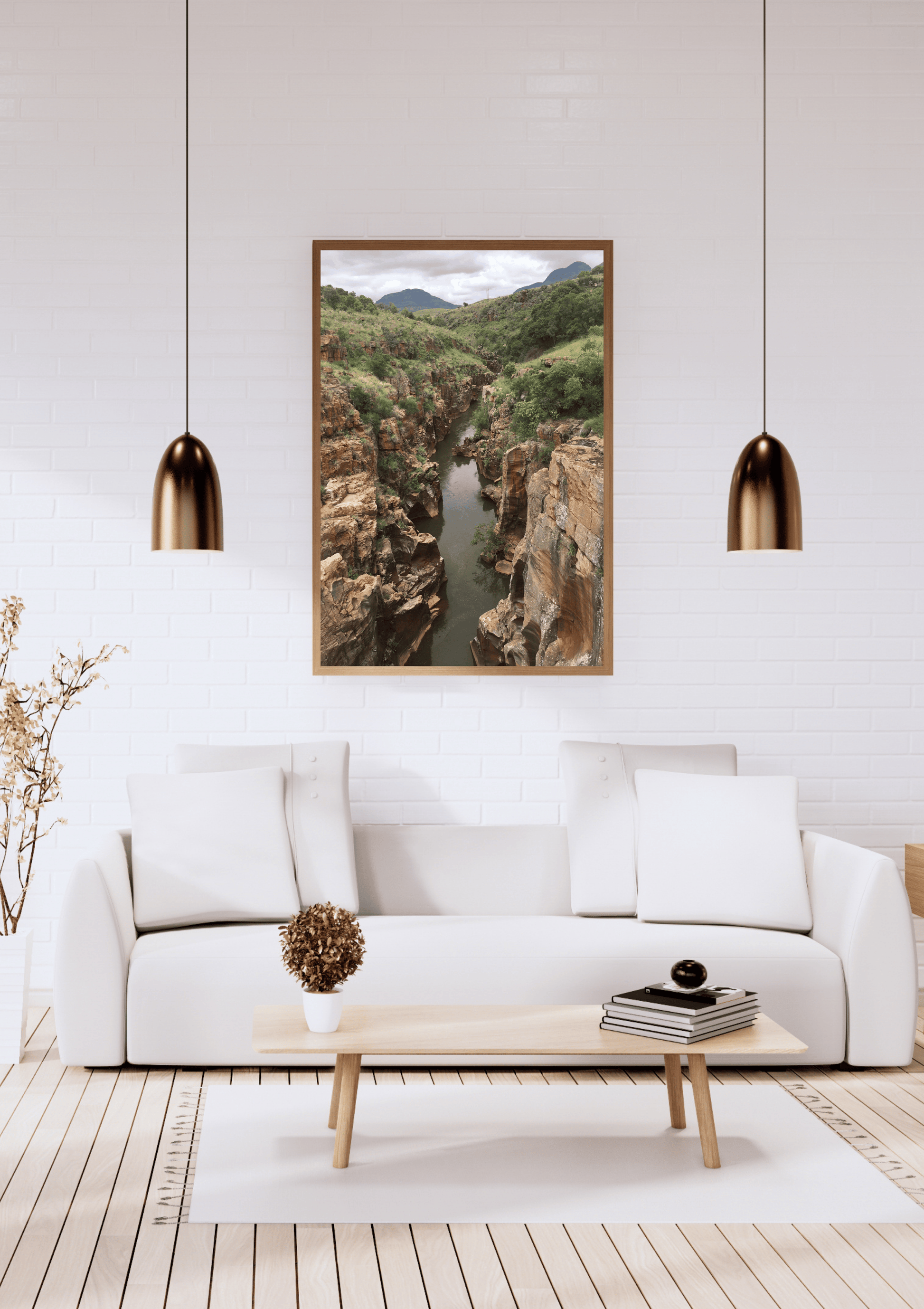 A framed landscape art print of a deep rocky canyon with a river flowing through it, displayed on a white brick wall above a modern white sofa in a styled living room.