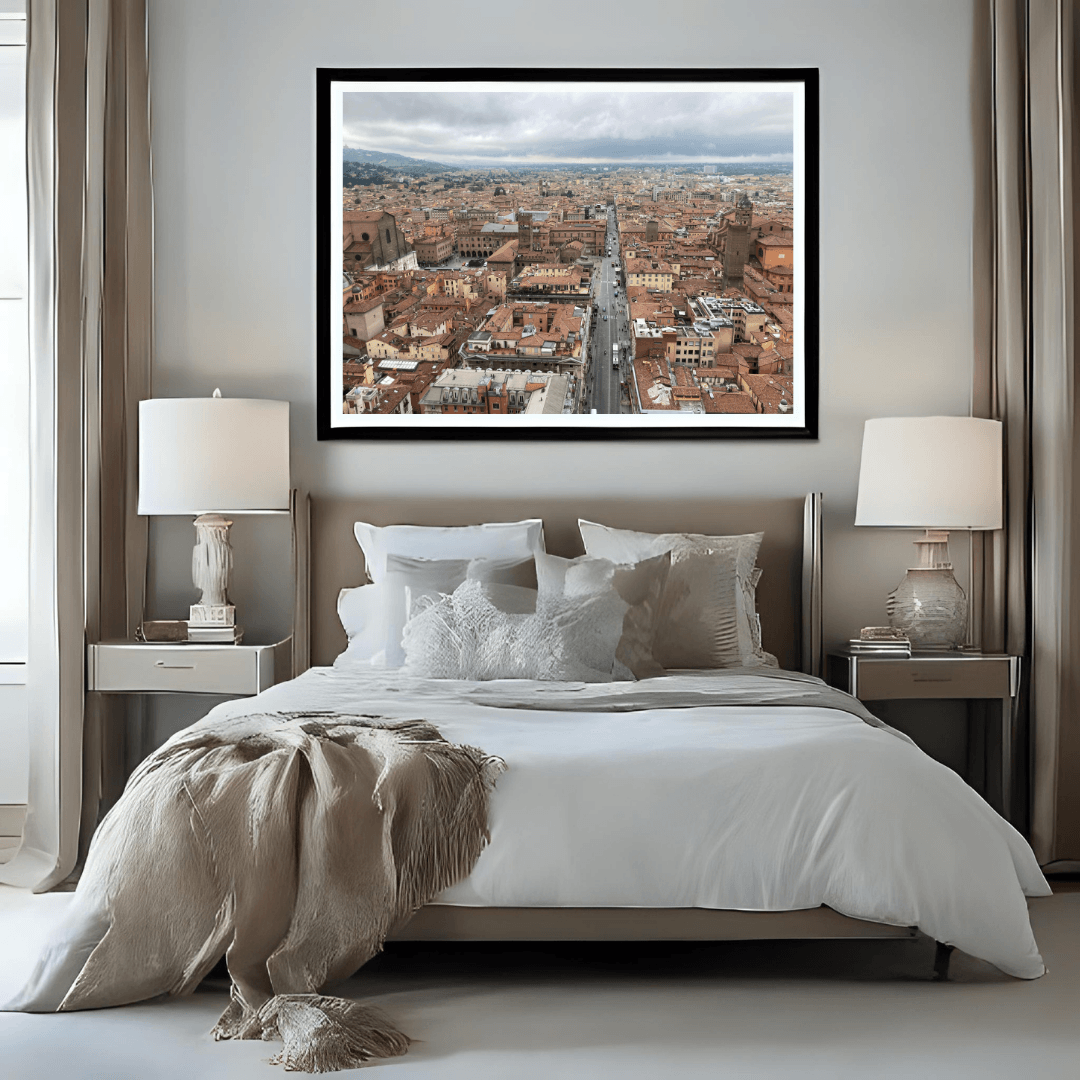 A framed panoramic cityscape print of Bologna's terracotta rooftops and a central street, displayed in a black frame on a light gray wall above a neatly made bed in a modern bedroom setting.