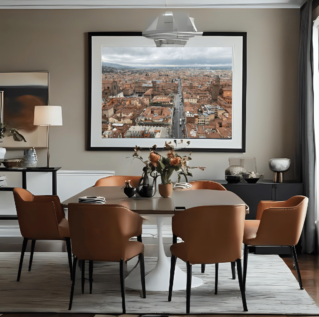 A large framed art print depicting a panoramic city view of Bologna hangs on a beige wall in a modern dining room setting with brown leather chairs and a white pedestal table.
