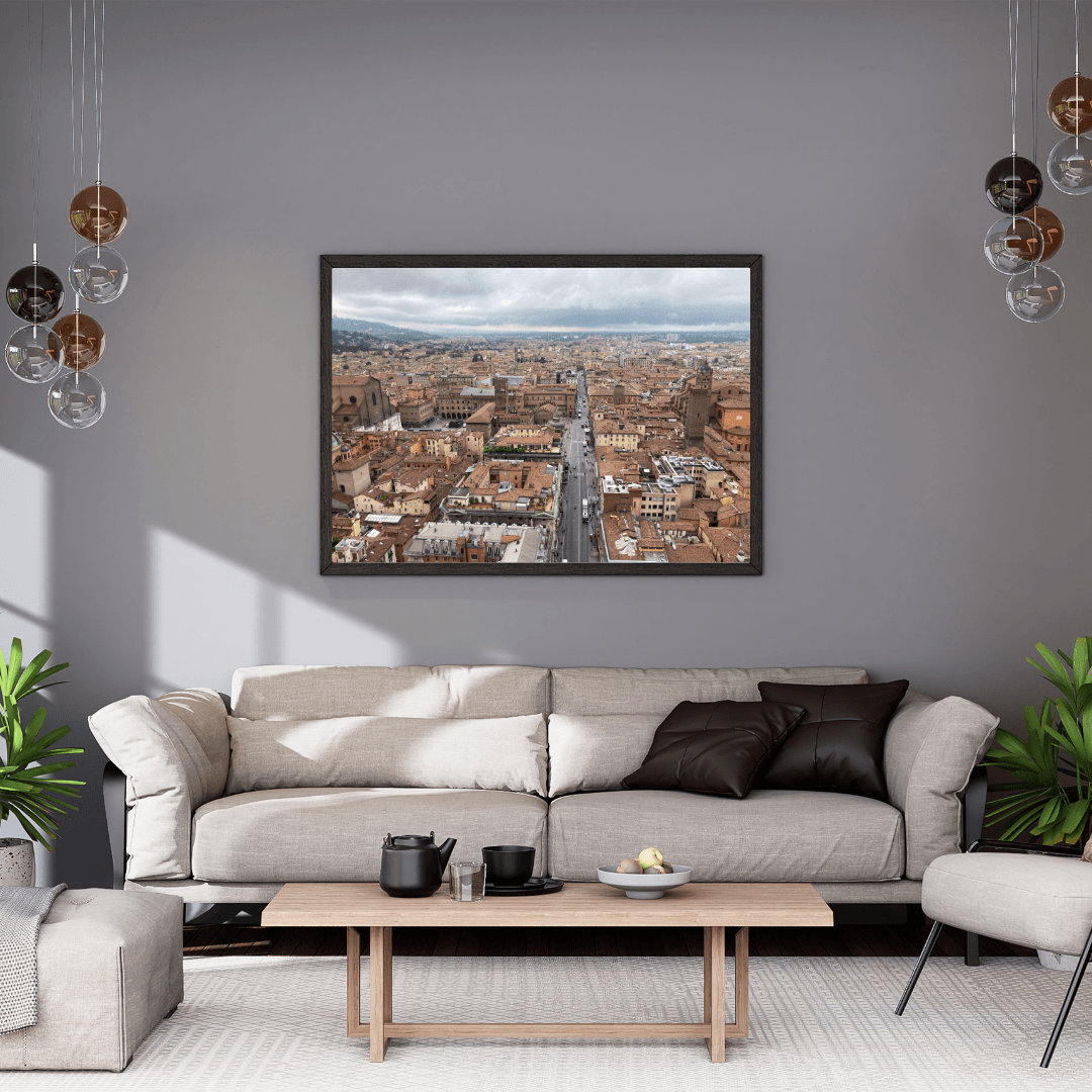A framed panoramic cityscape print of Bologna's terracotta rooftops and a long central street, displayed on a gray wall above a beige sofa in a modern living room.