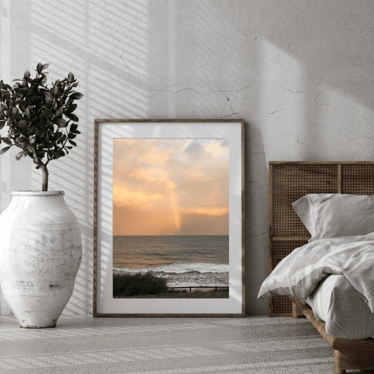 A framed vertical art print of a beach sunset with a rainbow over the ocean, styled in a modern bedroom setting next to a bed and a white vase with a plant.