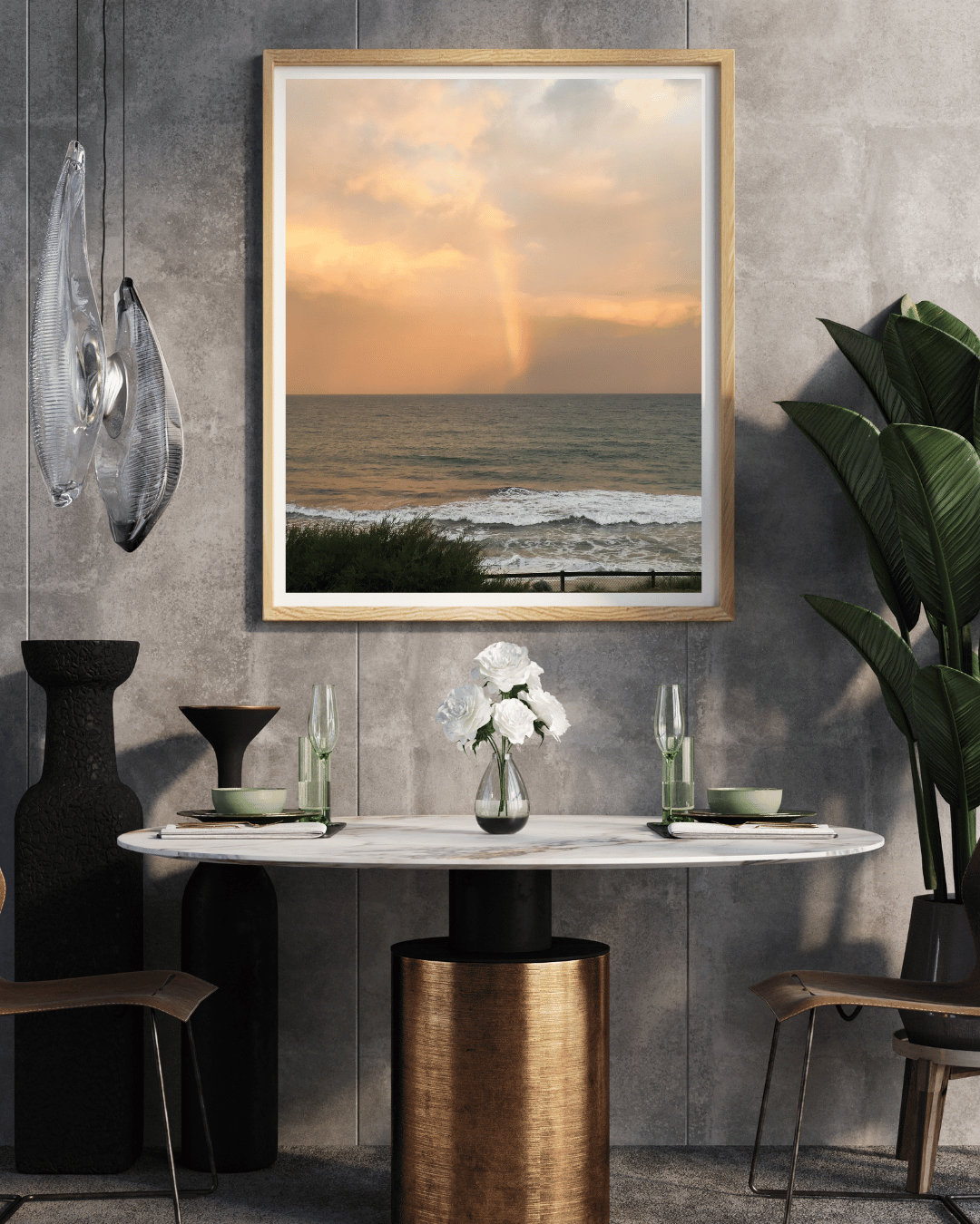 A framed vertical art print depicting a sunset over the ocean with a rainbow, styled in a modern dining room setting above a marble-top table.