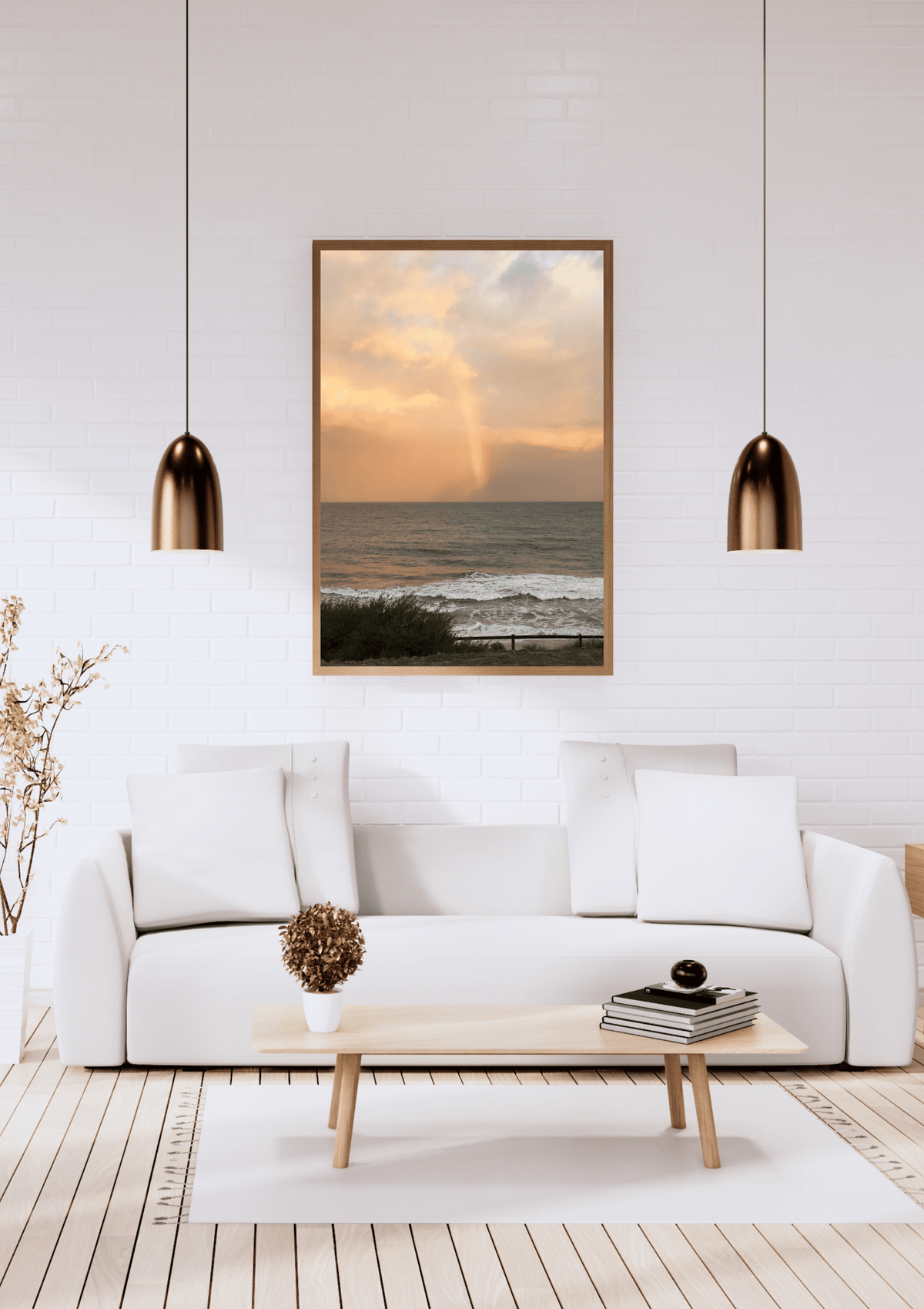 A framed vertical art print of a beach sunset with a rainbow is displayed on a white brick wall above a modern white sofa in a bright living room.