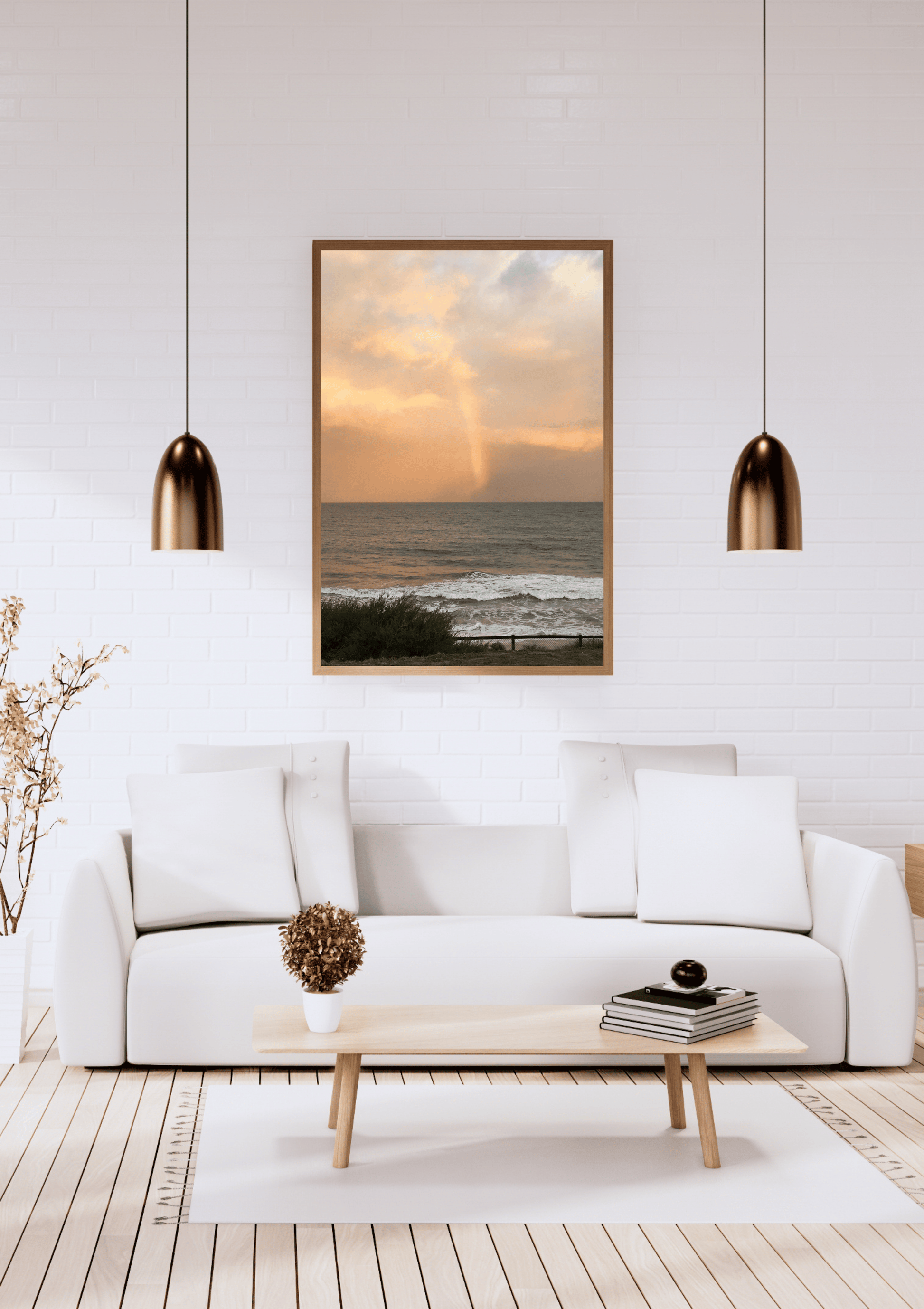 A framed vertical art print of a beach sunset with a rainbow is displayed on a white brick wall above a modern white sofa in a bright living room.