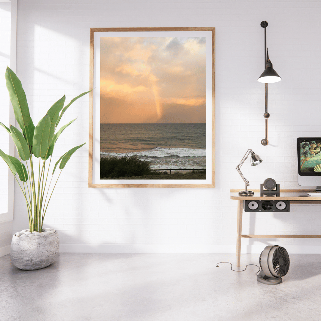 A framed vertical art print of an ocean sunset with a visible rainbow, displayed on a white wall in a modern home office setting with a desk, lamp, and potted plant.