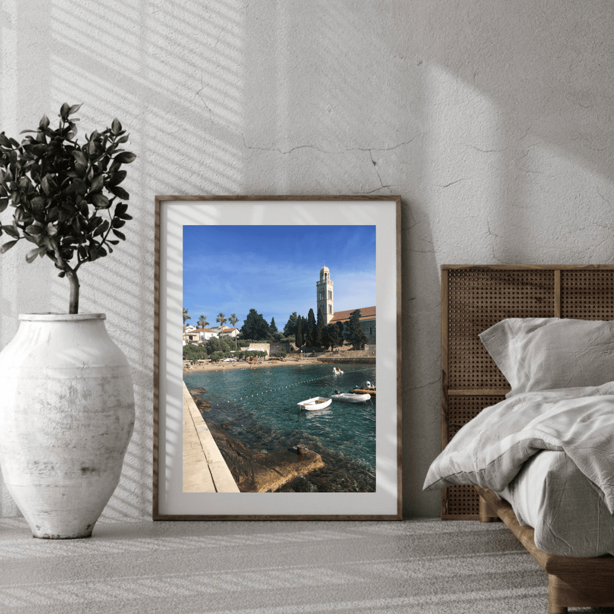 A framed art print depicting a scenic Croatian coastal harbor with boats and a tower under a blue sky, styled in a modern bedroom setting next to a bed and a large white vase.