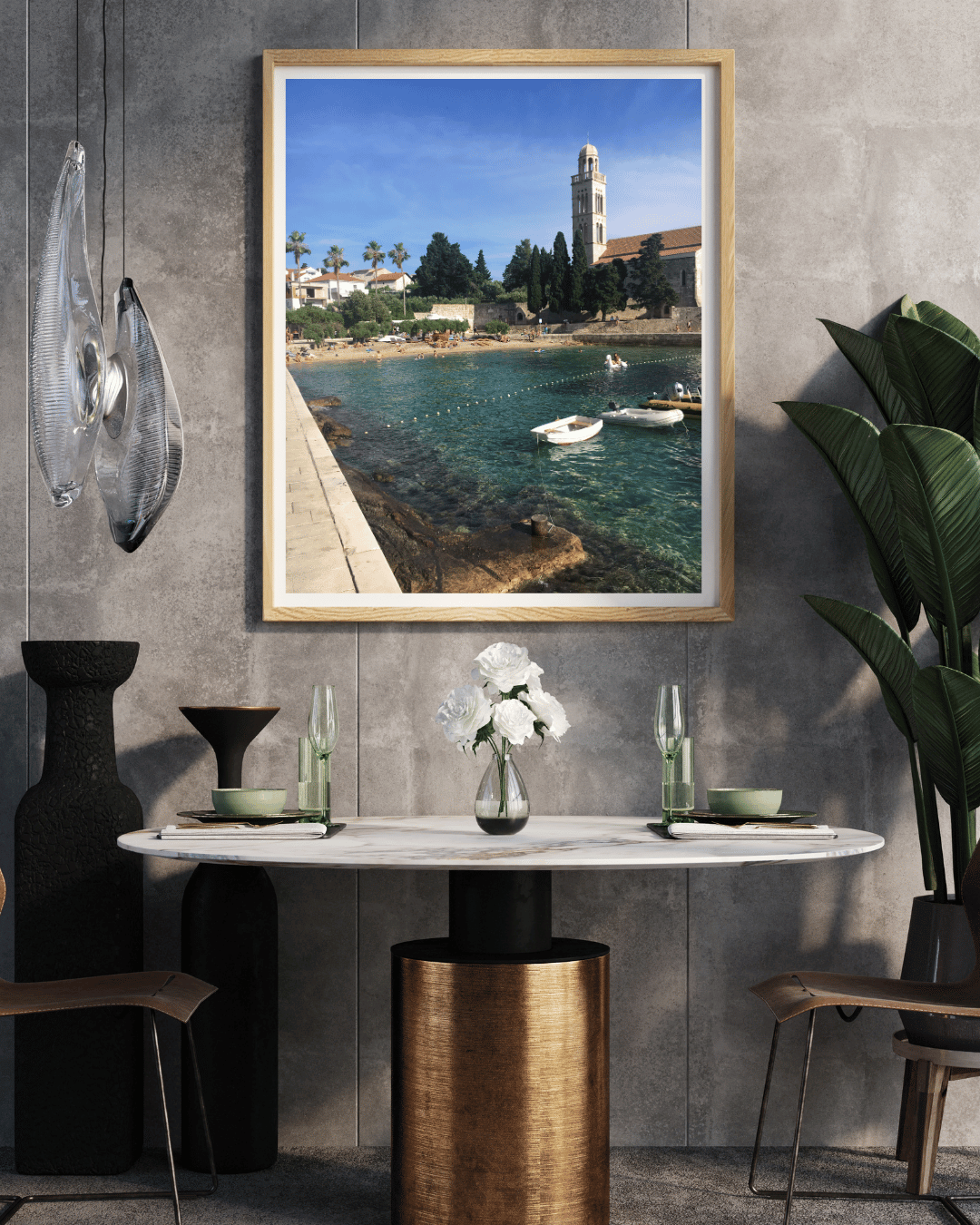 A framed coastal landscape print hangs on a textured gray wall above a dining table. The artwork shows a sunny Croatian seaside town with turquoise water, boats, trees, and a church tower.