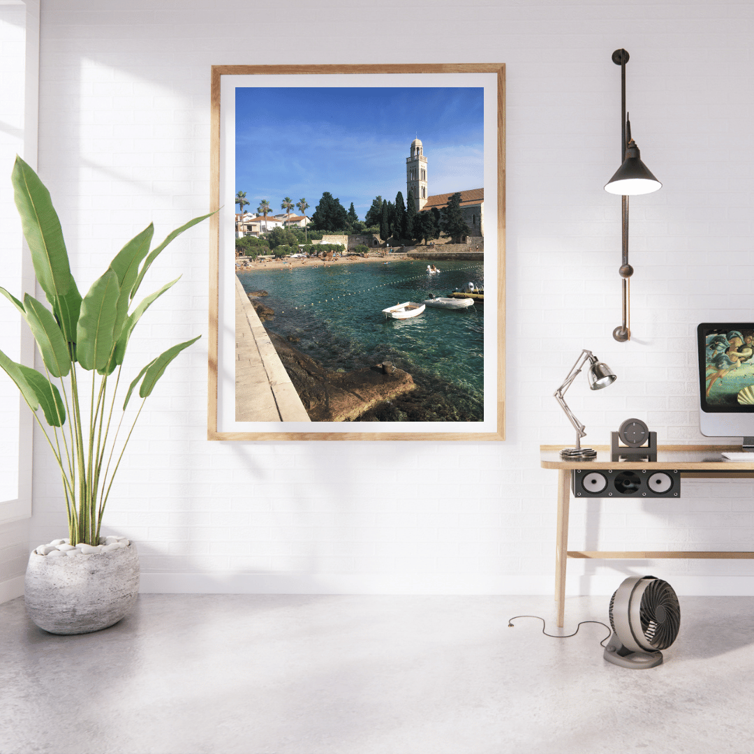 A framed art print depicting a scenic Croatian coastal harbor with boats and a tower under a blue sky, displayed on a white wall in a modern home office setting with a desk, lamp, and potted plant.