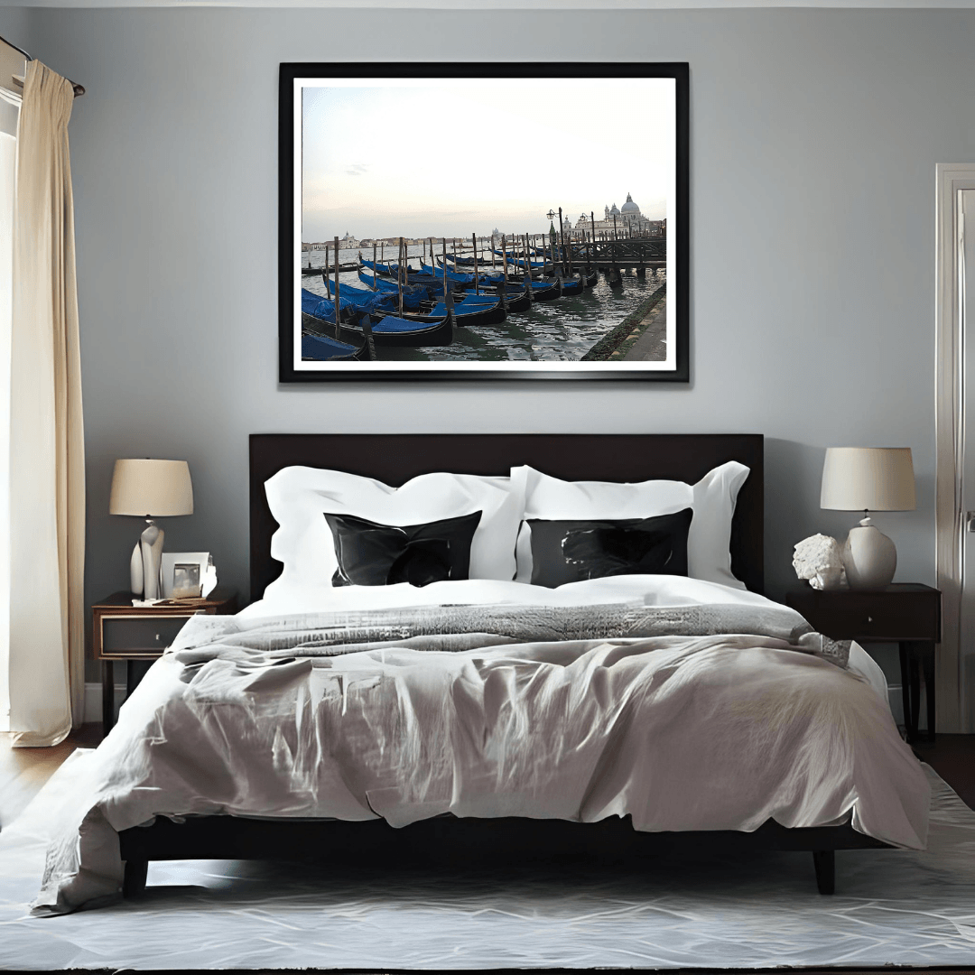 A framed photograph of blue gondolas in Venice, displayed in a black frame on a light gray wall above a neatly made bed in a modern bedroom setting.