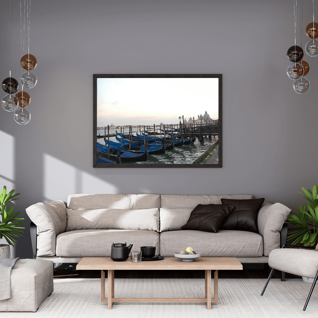 A framed art print depicting blue gondolas in Venice, displayed on a gray wall above a modern beige sofa in a styled living room.