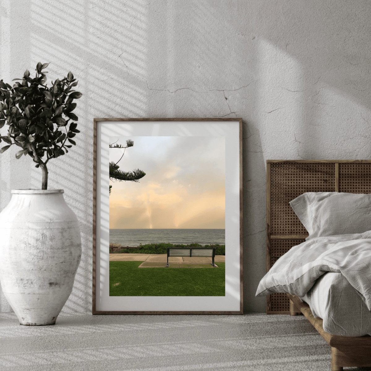 A framed vertical art print depicting a solitary bench overlooking a beach, styled in a modern bedroom setting next to a bed and a large white vase with a plant.