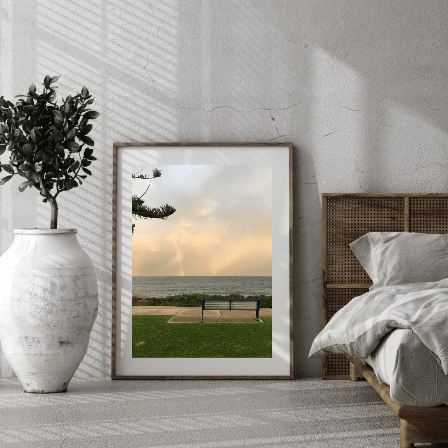 A framed vertical art print depicting a solitary bench overlooking a beach, styled in a modern bedroom setting next to a bed and a large white vase with a plant.