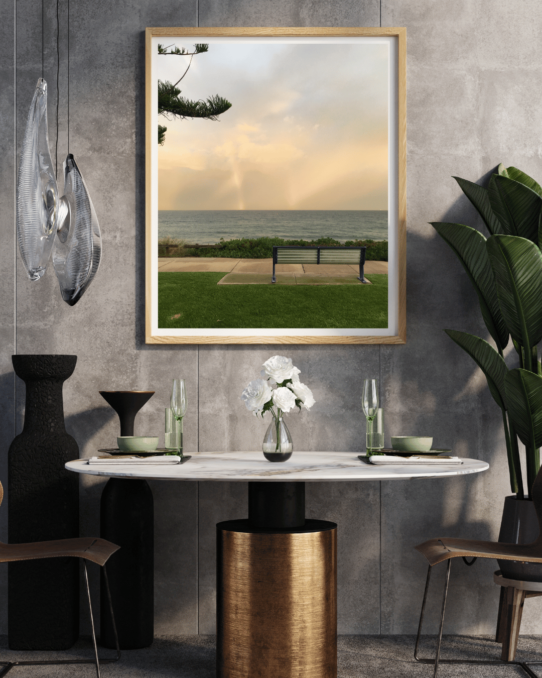A framed vertical art print depicting a  a solitary bench overlooking Cottesloe beach, styled on a gray concrete wall above a dining table in a modern interior.