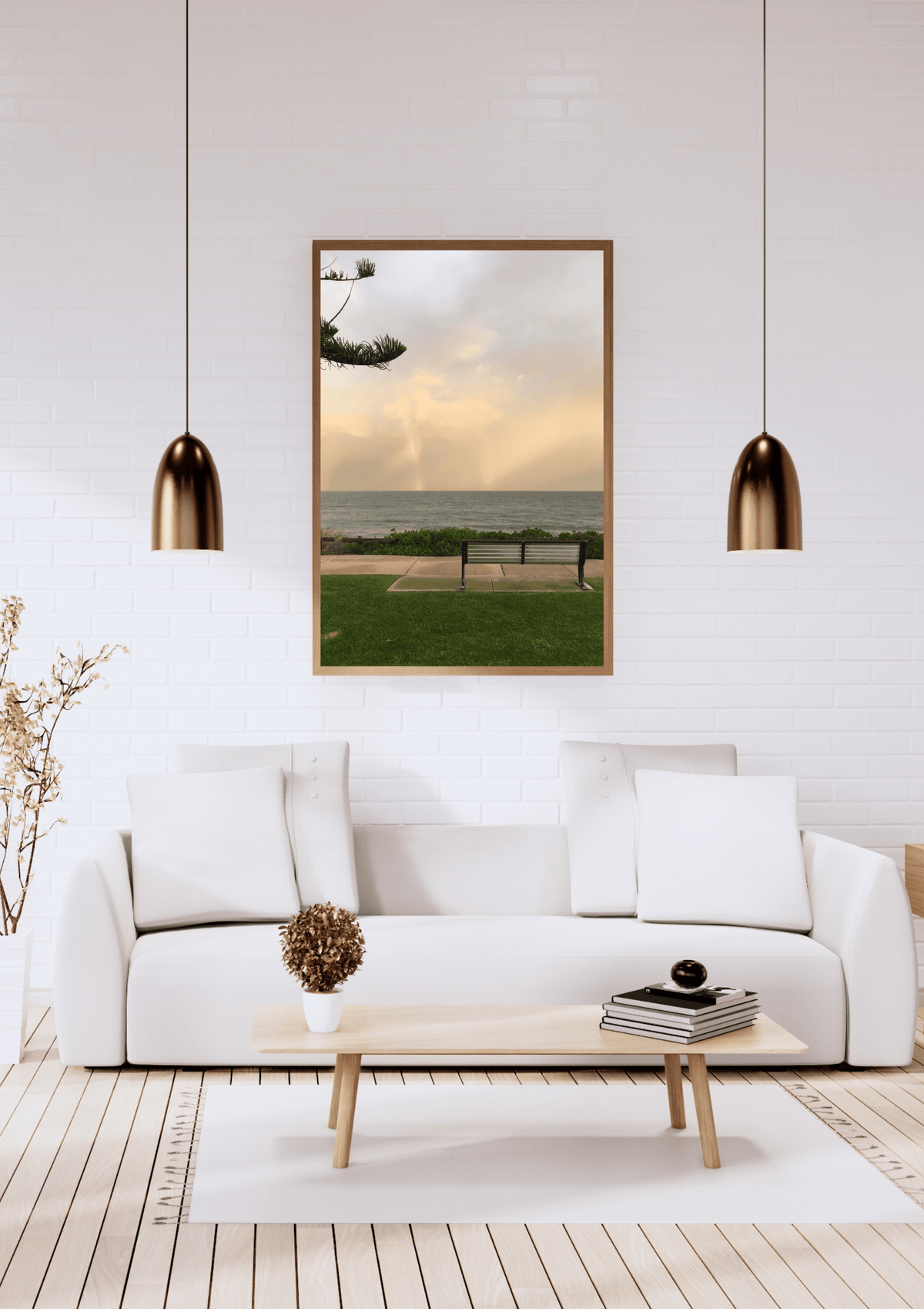 A framed vertical art print depicting a  a solitary bench overlooking Cottesloe beach, displayed on a white brick wall above a modern white sofa in a living room setting.