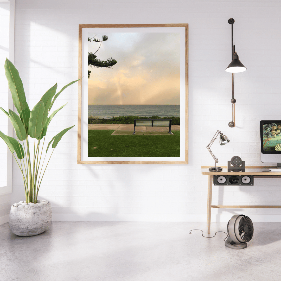 A framed vertical art print depicting a  a solitary bench overlooking Cottesloe beach, styled in a modern, sunlit room with a potted plant, desk, lamp, and computer.
