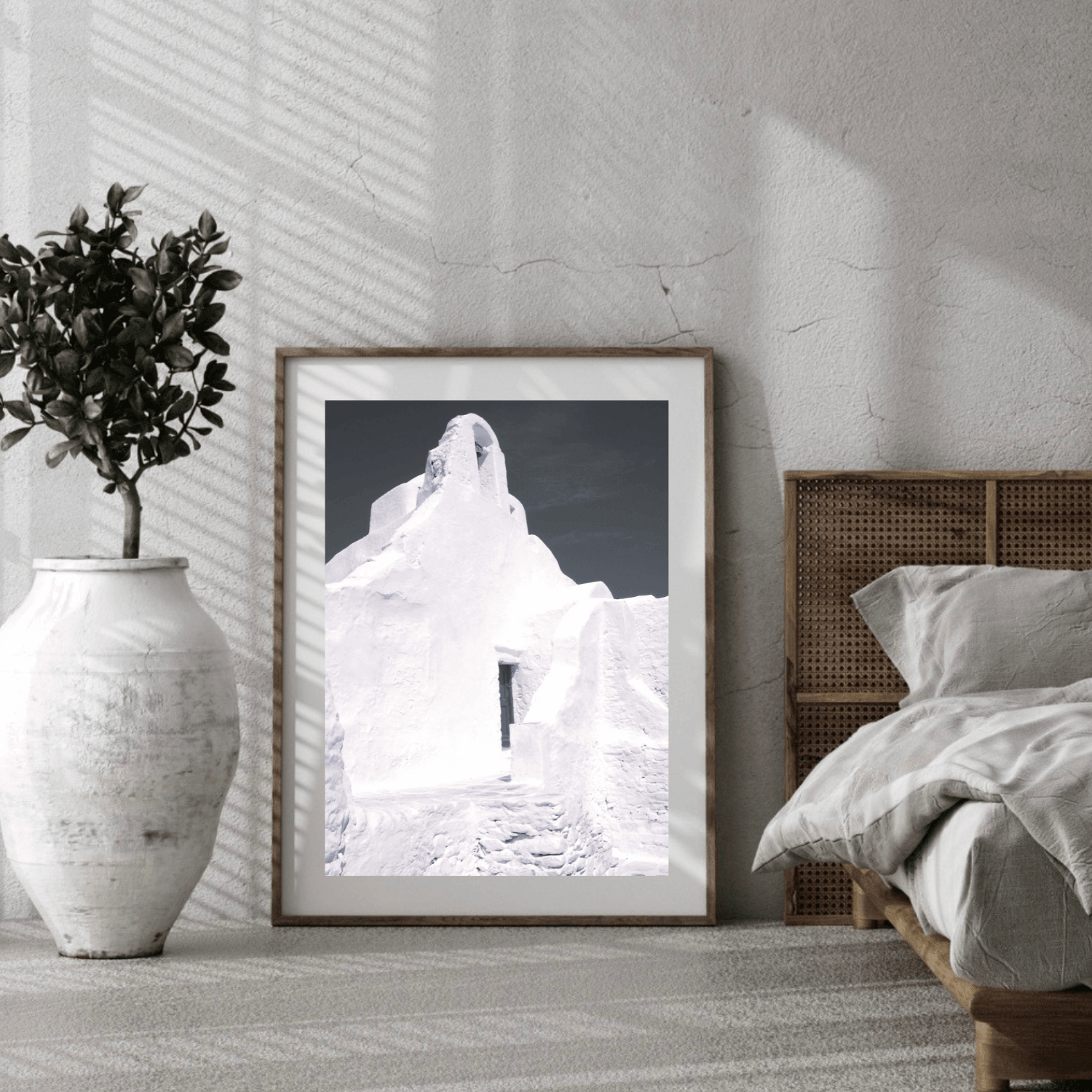 A framed black and white art print featuring a whitewashed building in Mykonos, Greece leaning against a textured white wall in a modern bedroom setting next to a bed and a white ceramic vase with a plant.