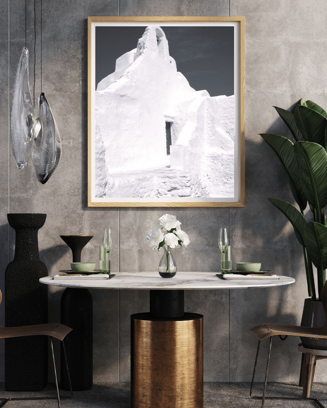 A framed black and white art print featuring a whitewashed building in Mykonos, Greece displayed on a gray concrete wall above a dining table in a modern styled room.