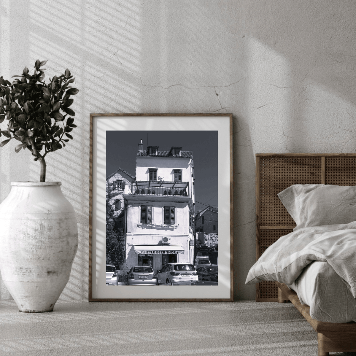A framed black and white architectural art print leaning against a textured wall in a styled bedroom setting next to a bed and a large white vase with a plant.