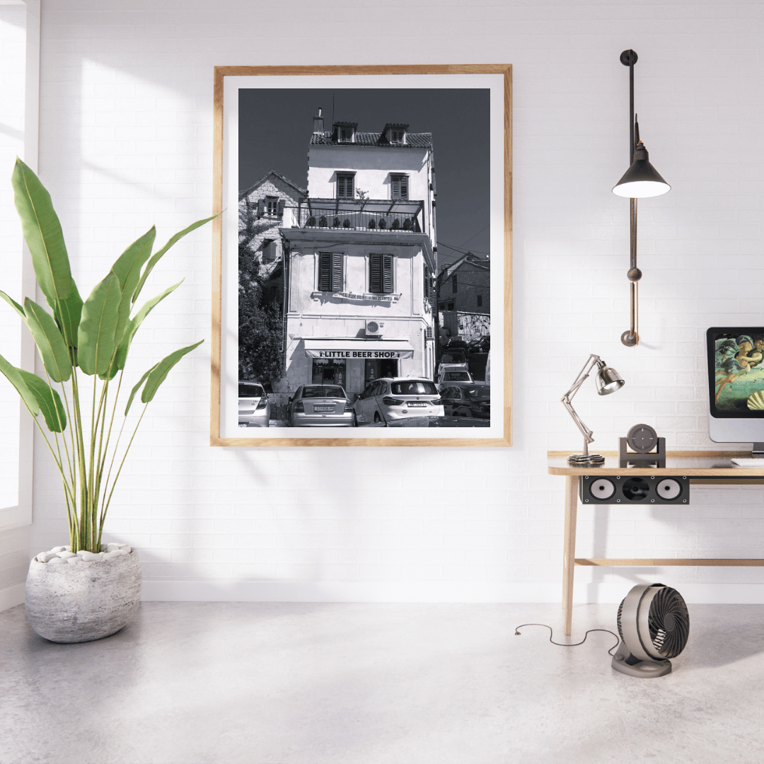 A framed black and white art print of a Parisian building is displayed on a white wall in a modern, sunlit room with a potted plant, desk, and lamp.