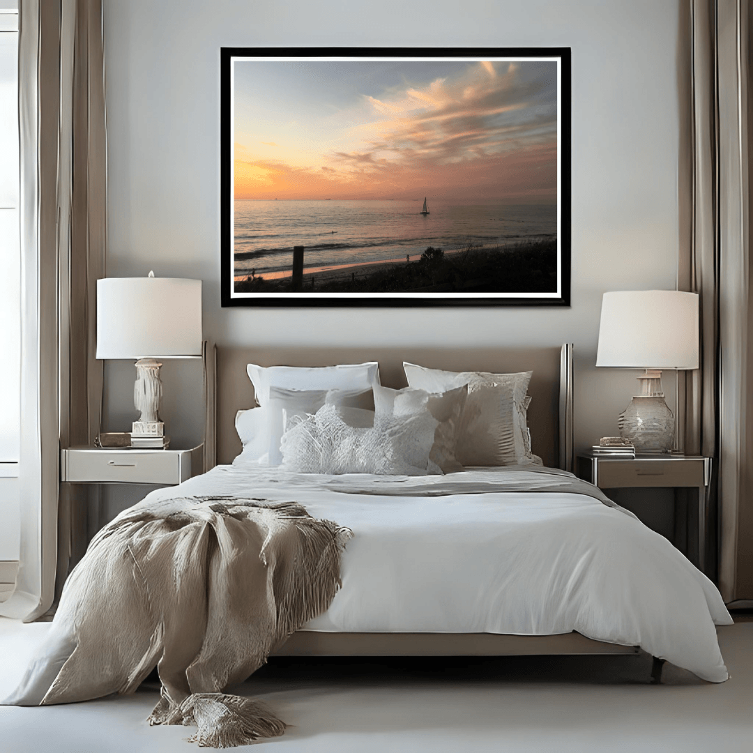 A framed wall art print of a solitary sailboat on the beach at sunset, displayed in a black frame above a neatly made bed in a modern, neutral-toned bedroom.