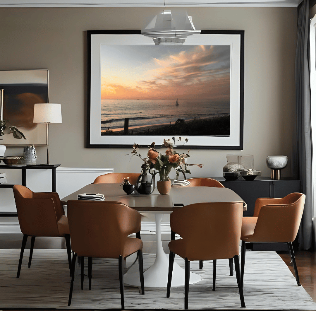 A large framed wall art print depicting a solitary sailboat on the beach at sunset displayed in a modern dining room setting above a dining table and six brown upholstered chairs.