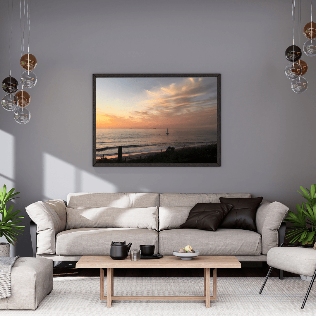 A framed horizontal landscape print of a solitary sailboat on the beach at sunset, displayed on a gray wall above a modern beige sofa in a styled living room.