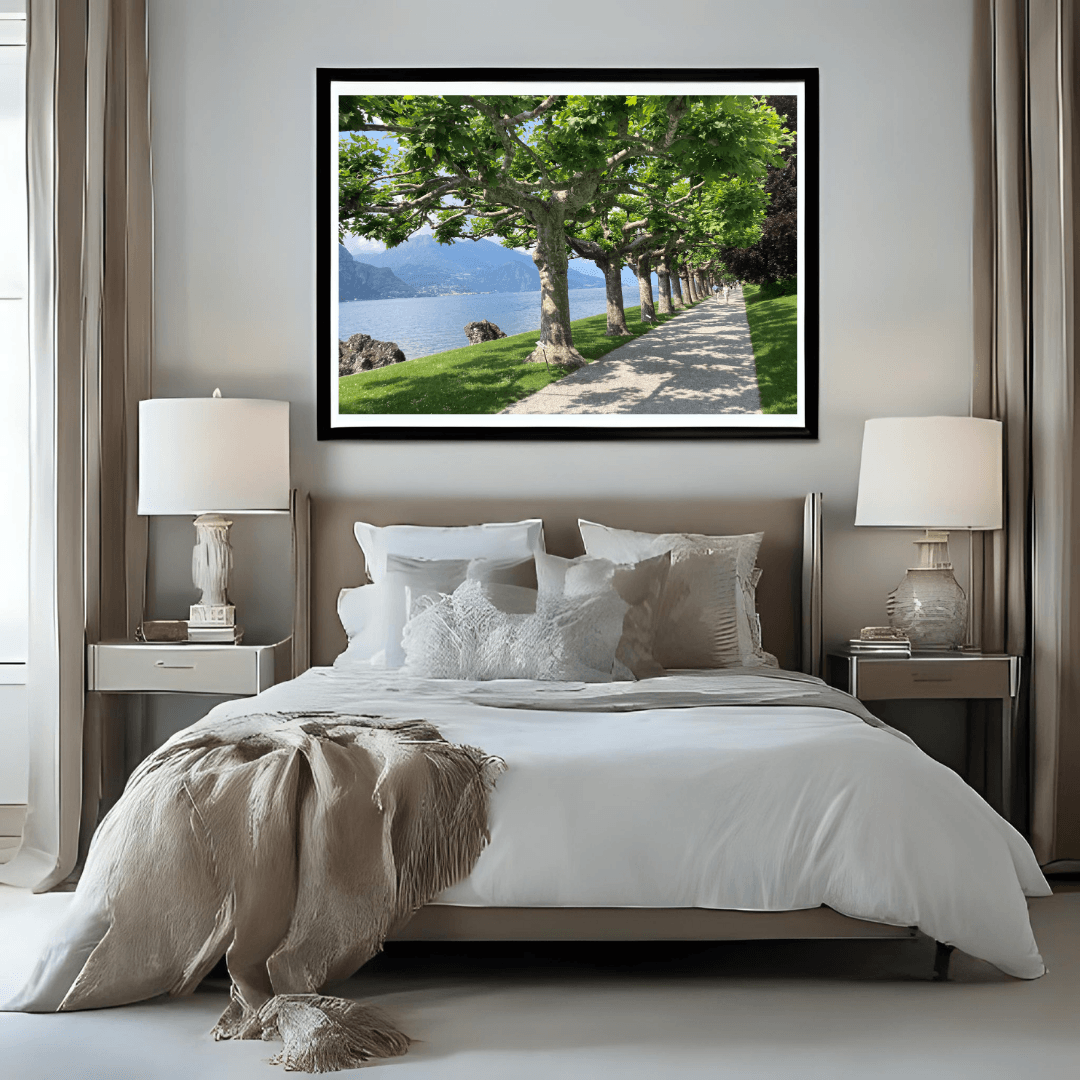 A framed landscape art print of a scenic Lake Como path lined with green trees, displayed on a light gray wall above a neatly made bed in a modern bedroom setting.