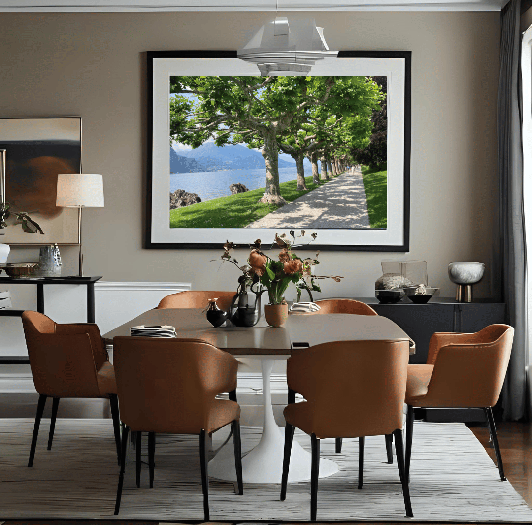 A large framed landscape art print depicting a scenic tree-lined path by  Lake Como, displayed on a beige wall in a modern dining room setting with a dining table and six brown upholstered chairs.