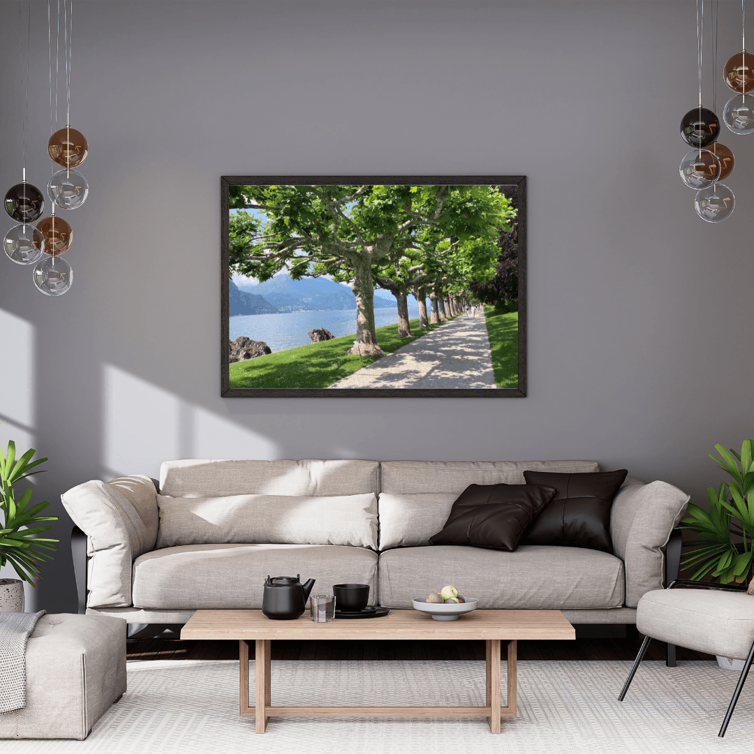 A framed landscape art print depicting a scenic  Lake Como path lined with green trees next to the lake, displayed on a gray wall above a modern beige sofa in a styled living room.