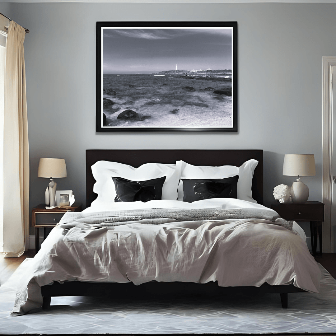 A black-framed black and white coastal landscape print hangs above a bed in a modern bedroom with gray walls, white bedding, and dark wood furniture.