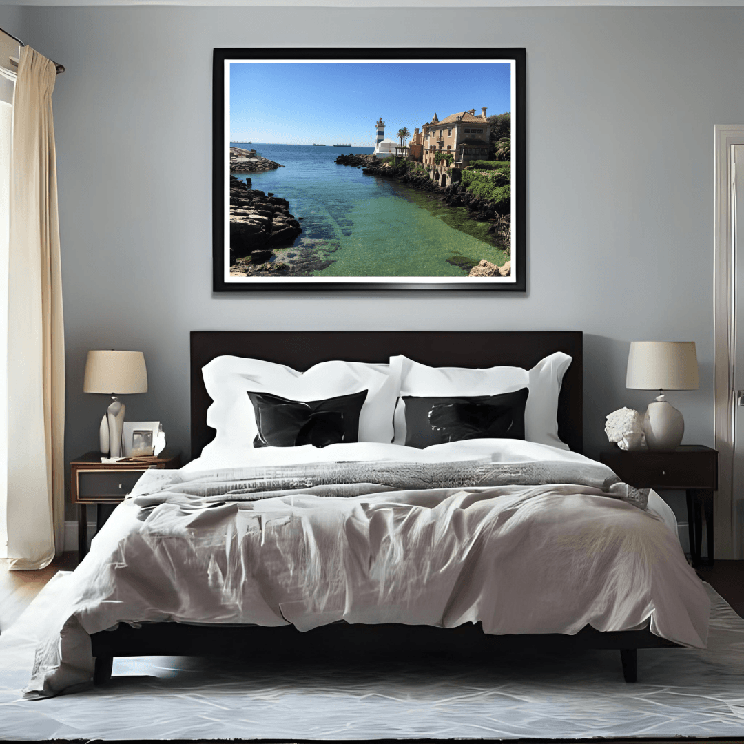 A framed coastal landscape print hangs above a bed in a modern bedroom. The artwork shows a sunny seaside town in Cascais with turquoise water, rocky shoreline, and buildings under a blue sky.