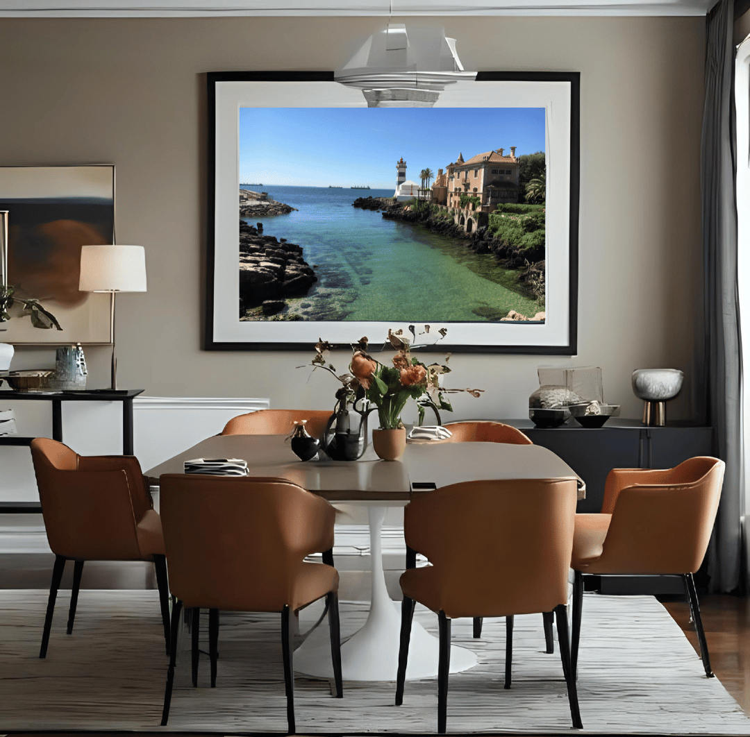A large framed wall art print depicting a coastal seaside town of Cascais with turquoise water and buildings, displayed above a dining table with brown chairs in a modern living room setting.