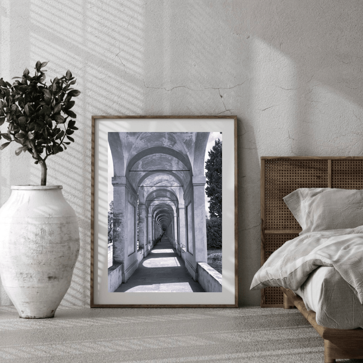 A framed black and white architectural art print leaning against a textured wall in a modern bedroom setting, next to a bed and a large white vase with a potted plant.