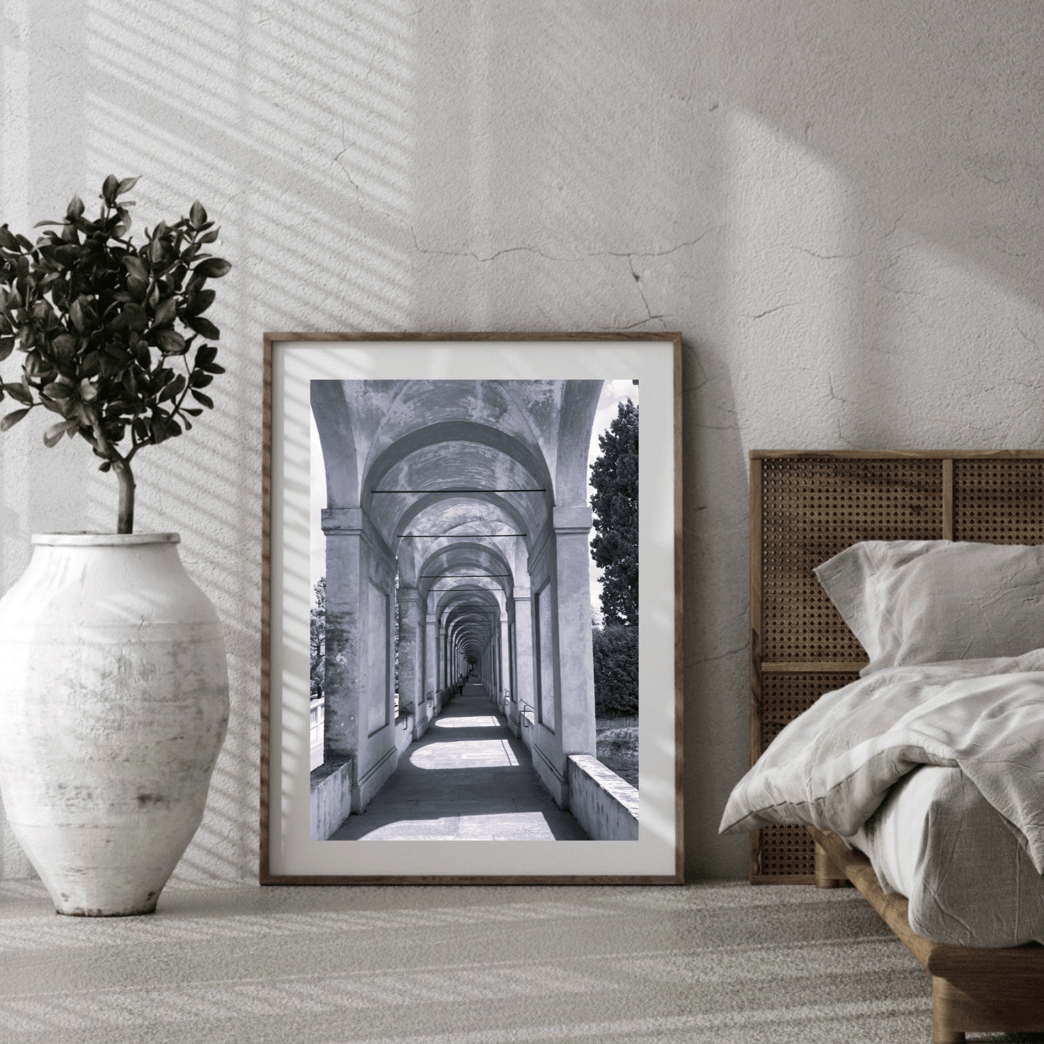 A framed black and white architectural art print leaning against a textured wall in a modern bedroom setting, next to a bed and a large white vase with a potted plant.