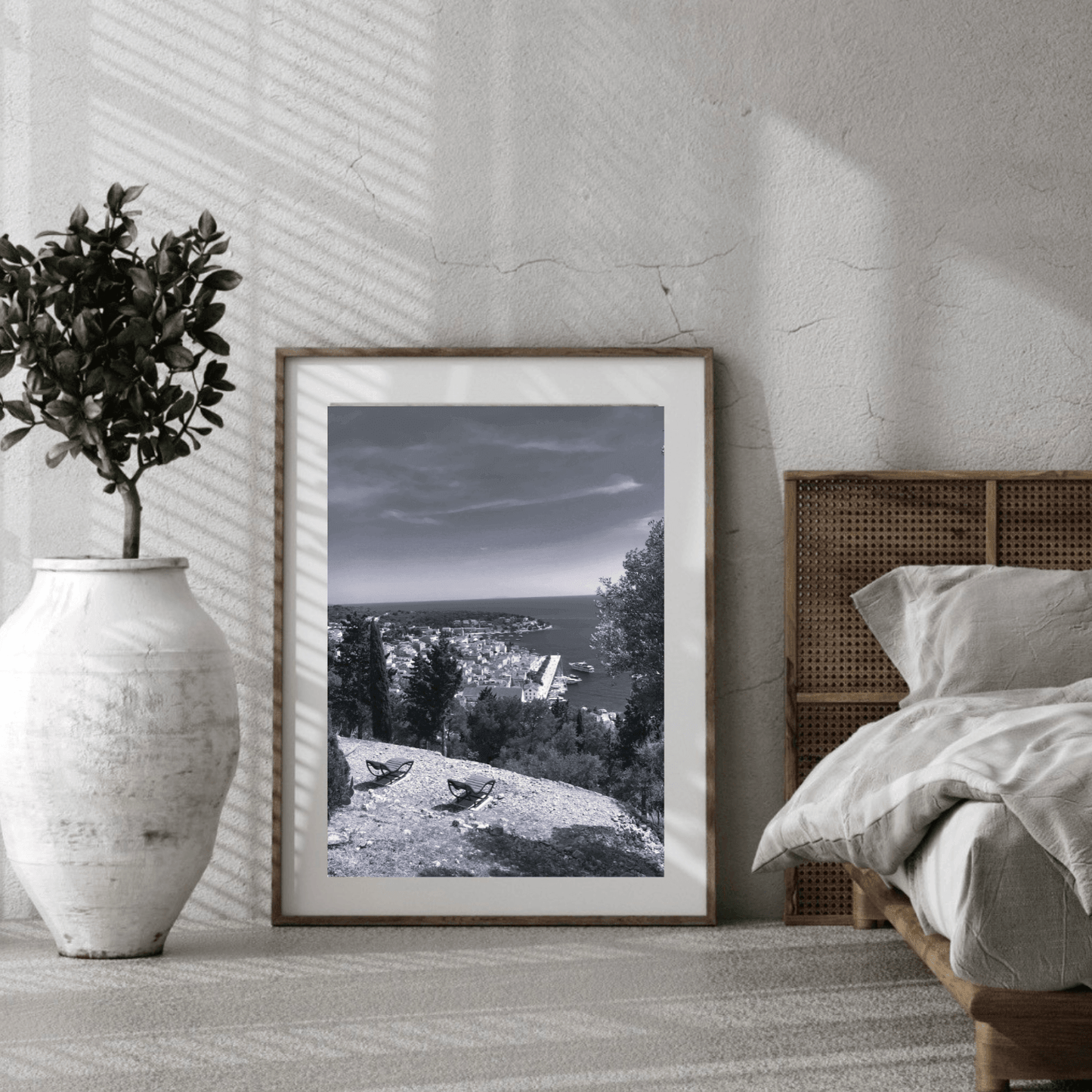 A framed black and white landscape photograph leaning against a textured wall in a modern bedroom setting, next to a bed and a large white vase with a potted plant.