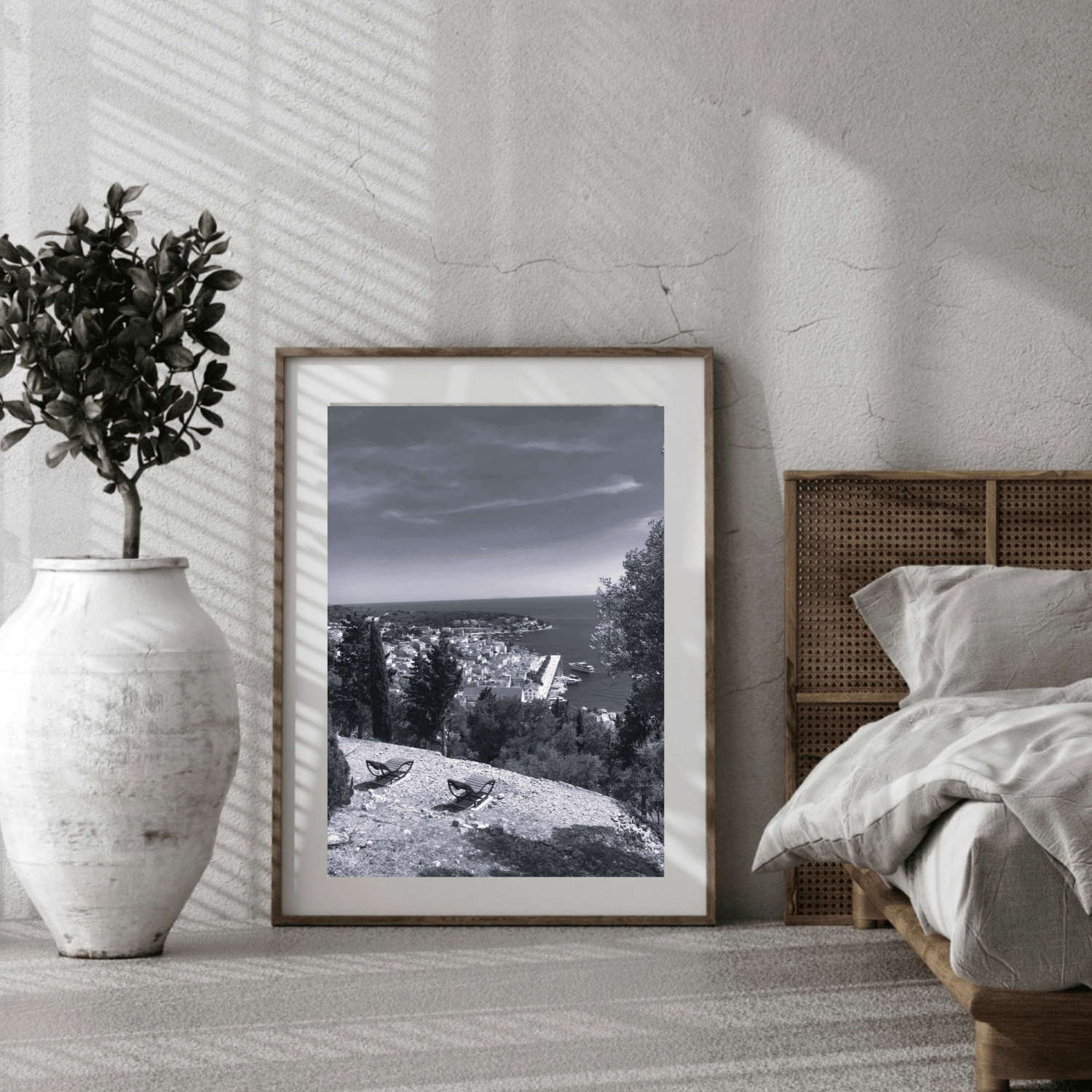 A framed black and white landscape photograph leaning against a textured wall in a modern bedroom setting, next to a bed and a large white vase with a potted plant.